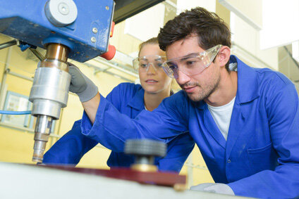 Jetzt bewerben | © fotolia Zwei Auszubildende, eine junge Frau links und ein junger Mann rechts, beide mit Blaumann-Overall gekleidet und mit Schutzbrillen, arbeiten an einer Standbohrmaschine, im Hintergrund Werkstatt | © fotolia